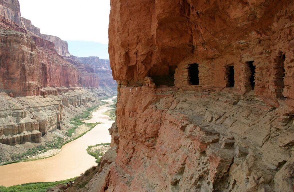 En el lado derecho de la imagen hay una gruesa pared carmesí con cuevas alineadas y talladas en la pared del cañón. A la izquierda hay otra pared de color naranja pálido, que se eleva sobre el fangoso río Colorado.