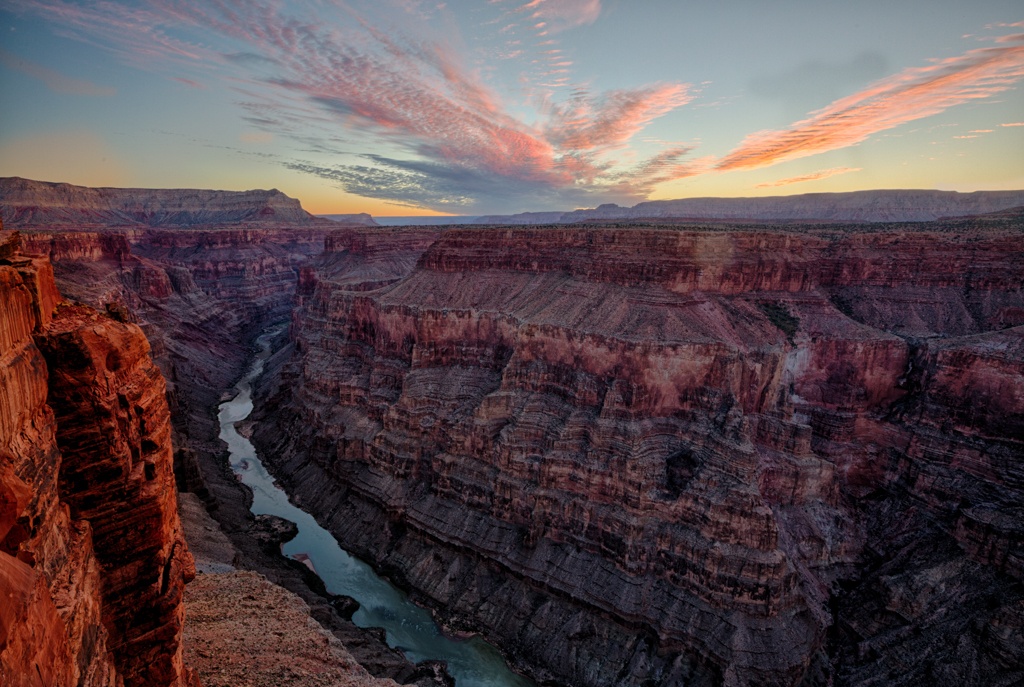 火熱的太陽從峽谷上空落下，粉紅色和紫色的雲彩引人注目，紫色和紅色的岩石層層排列在下面的綠松石河上