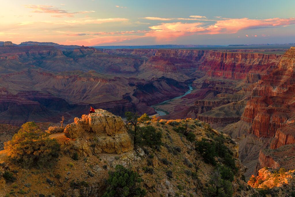 屏幕外的日落将高耸的岩石染成黄色，一个人坐在上面，望向远方。 峡谷是一系列紫色和橙色延伸到地平线，并由一条绚丽的蓝色科罗拉多河锚定